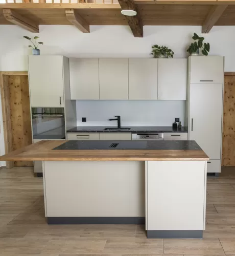 Moderne Küche mit heller Einbauküche, Kochinsel mit Holzarbeitsplatte und integrierten Geräten in einem offenen Wohnraum mit sichtbaren Holzbalken.