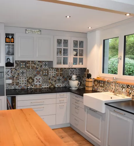 Individuell gestaltete Küche mit hellen Fronten, integrierten Einbaugeräten und grosszügiger Kochinsel mit massiver Holzarbeitsplatte. Der dekorative Fliesenspiegel setzt einen farbigen Akzent und bildet einen Kontrast zu den klaren Linien der Möblierung. Warme Holzböden und die offene Raumgestaltung schaffen eine einladende und wohnliche Atmosphäre.