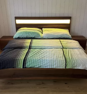 Ein modernes Doppelbett aus dunkel gemasertem Holz steht im Mittelpunkt des Schlafzimmers. Das Kopfteil ist mit einem horizontalen, warm leuchtenden Lichtstreifen versehen, der den Raum sanft ausleuchtet. Auf dem Bett liegt eine Bettwäsche in harmonischen Grün-, Blau- und Weißtönen mit einem feinen Linienmuster. Links und rechts befinden sich passende Nachttische aus dem gleichen Holz. Der Raum hat einen hellen Holzboden sowie weiss vertäfelte Wände, die eine ruhige, natürliche Atmosphäre schaffen.
