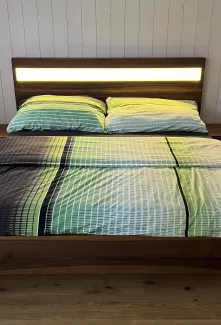 Ein modernes Doppelbett aus dunkel gemasertem Holz steht im Mittelpunkt des Schlafzimmers. Das Kopfteil ist mit einem horizontalen, warm leuchtenden Lichtstreifen versehen, der den Raum sanft ausleuchtet. Auf dem Bett liegt eine Bettwäsche in harmonischen Grün-, Blau- und Weißtönen mit einem feinen Linienmuster. Links und rechts befinden sich passende Nachttische aus dem gleichen Holz. Der Raum hat einen hellen Holzboden sowie weiss vertäfelte Wände, die eine ruhige, natürliche Atmosphäre schaffen.