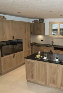 Massgefertigte Küche aus warmem Eichenholz mit zentraler Kochinsel und dunkler Arbeitsplatte. Die Kombination aus natürlichen Holzfronten, klaren Linien und modernen Einbaugeräten schafft eine zeitlose und funktionale Raumgestaltung. Grosszügige Fenster sorgen für viel Tageslicht und unterstreichen die helle, freundliche Wohnatmosphäre.