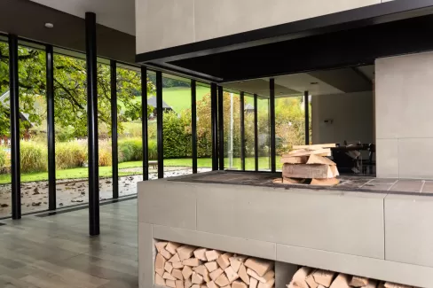 Moderner Essbereich mit grosser Glasfront und Blick in den Garten. In der Mitte steht ein runder Holztisch mit schwarzen Stühlen auf einem hellen Holzboden. Das natürliche Licht schafft eine offene, freundliche Atmosphäre.