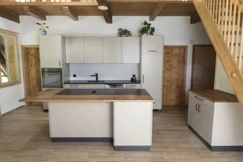 Moderne Küche mit heller Einbauküche, Kochinsel mit Holzarbeitsplatte und integrierten Geräten in einem offenen Wohnraum mit sichtbaren Holzbalken.