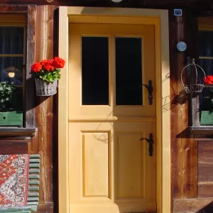 Helle Holztür mit zwei Glaseinsätzen in der oberen Hälfte, eingebaut in eine rustikale Hausfassade aus dunklem Holz. Neben der Tür hängen Blumenkörbe mit roten Geranien, und vor dem Eingang liegt eine Fussmatte – traditioneller Schweizer Hausstil.