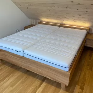 Ein modernes Doppelbett aus dunkel gemasertem Holz steht im Mittelpunkt des Schlafzimmers. Das Kopfteil ist mit einem horizontalen, warm leuchtenden Lichtstreifen versehen, der den Raum sanft ausleuchtet. Auf dem Bett liegt eine Bettwäsche in harmonischen Grün-, Blau- und Weißtönen mit einem feinen Linienmuster. Links und rechts befinden sich passende Nachttische aus dem gleichen Holz. Der Raum hat einen hellen Holzboden sowie weiss vertäfelte Wände, die eine ruhige, natürliche Atmosphäre schaffen.