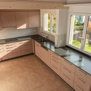 Moderne Küche mit matten, roséfarbenen Fronten, Edelstahlgriffen und hellbraunem Fliesenboden. Die Küche hat eine L-Form, eine dunkle Arbeitsplatte und eine Holzdecke mit natürlicher Maserung.