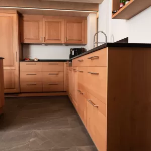 Innenansicht einer grosszügigen Küche mit massgefertigten Holzfronten und dunkler Arbeitsfläche. In der Mitte befindet sich eine Kochinsel mit integriertem Kochfeld. Einbauschränke, Backofen und Spüle sind entlang der Wände angeordnet. Die Kombination aus Holzdecke, Holzmöbeln und Steinboden verleiht dem Raum eine hochwertige, warme Atmosphäre.