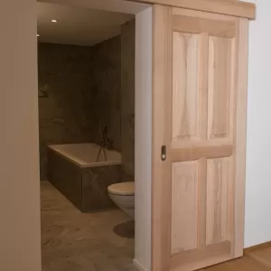 Massgefertigte Schiebetür aus Schweizer Eschenholz in heller, natürlicher Optik. Die Tür öffnet den Blick in ein modern gestaltetes Badezimmer mit grossformatigen Steinplatten, einer integrierten Badewanne und einer zeitlosen, schlichten Ausstattung. Die Kombination aus warmem Holz und reduzierten Materialien schafft eine ruhige, hochwertige Wohnatmosphäre.