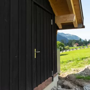 Aussenansicht eines Holzhauses mit dunkel lasierter Fassade, Eingangstür und Fenster mit grünen Holzläden unter einem grossen Dachvorsprung