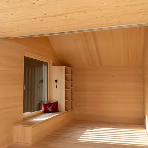 Holzverkleidete Sitznische am Fenster mit integrierten Schubladen, offenen Regalen und roten Zierkissen in einem hellen Innenraum.