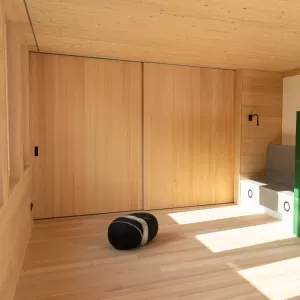 Heller Innenraum mit durchgehendem Holzausbau, Einbauschrank, Sitznische am Fenster und modernem grünen Kaminofen.