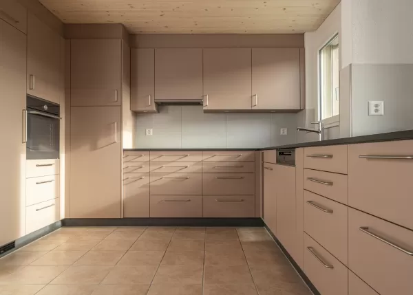 Moderne Küche mit matten, roséfarbenen Fronten, Edelstahlgriffen und hellbraunem Fliesenboden. Die Küche hat eine L-Form, eine dunkle Arbeitsplatte und eine Holzdecke mit natürlicher Maserung.