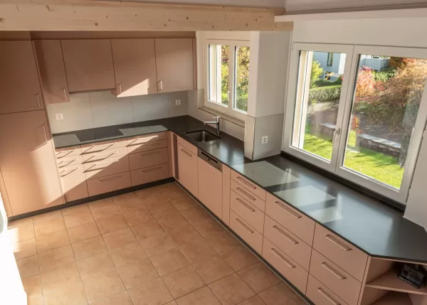 Moderne Küche mit matten, roséfarbenen Fronten, Edelstahlgriffen und hellbraunem Fliesenboden. Die Küche hat eine L-Form, eine dunkle Arbeitsplatte und eine Holzdecke mit natürlicher Maserung.