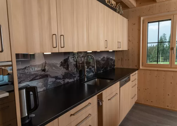 Innenansicht einer hochwertigen Küche in Holzbauweise mit hellen Holzfronten und sichtbarer Holzbalkendecke. Die Küche verfügt über eine dunkle Arbeitsfläche, eine grosse Kochinsel und eine Rückwand mit Bergmotiv. Ein Holzfenster sorgt für Tageslicht und unterstreicht die warme, alpine Atmosphäre.