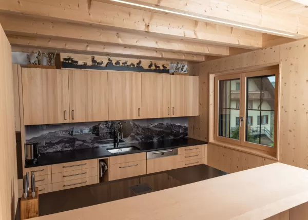 Innenansicht einer hochwertigen Küche in Holzbauweise mit hellen Holzfronten und sichtbarer Holzbalkendecke. Die Küche verfügt über eine dunkle Arbeitsfläche, eine grosse Kochinsel und eine Rückwand mit Bergmotiv. Ein Holzfenster sorgt für Tageslicht und unterstreicht die warme, alpine Atmosphäre.