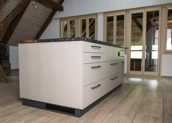 Moderne Küche mit heller Einbauküche, Kochinsel mit Holzarbeitsplatte und integrierten Geräten in einem offenen Wohnraum mit sichtbaren Holzbalken.