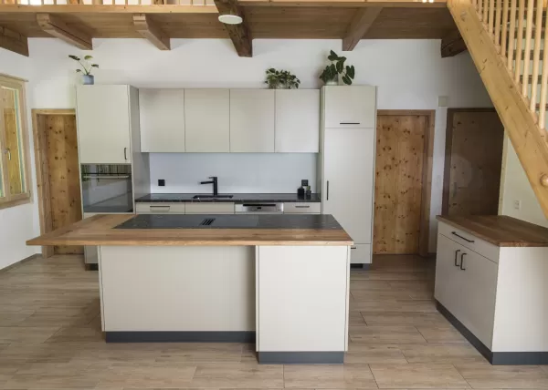 Moderne Küche mit heller Einbauküche, Kochinsel mit Holzarbeitsplatte und integrierten Geräten in einem offenen Wohnraum mit sichtbaren Holzbalken.