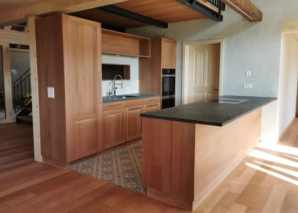 Moderne Küche mit massgefertigten Holzschränken, Kücheninsel mit dunkler Arbeitsplatte und warmem Parkettboden in einem hellen Innenraum.