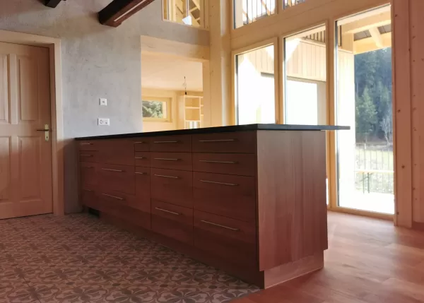 Moderne Küche mit massgefertigten Holzschränken, Kücheninsel mit dunkler Arbeitsplatte und warmem Parkettboden in einem hellen Innenraum.