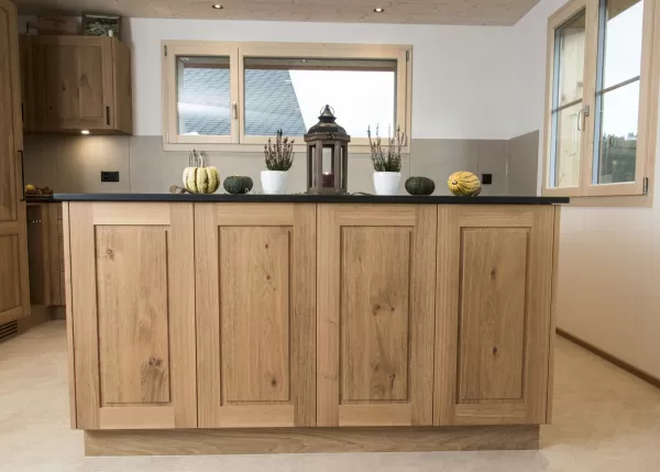 Massgefertigte Küche aus warmem Eichenholz mit zentraler Kochinsel und dunkler Arbeitsplatte. Die Kombination aus natürlichen Holzfronten, klaren Linien und modernen Einbaugeräten schafft eine zeitlose und funktionale Raumgestaltung. Grosszügige Fenster sorgen für viel Tageslicht und unterstreichen die helle, freundliche Wohnatmosphäre.