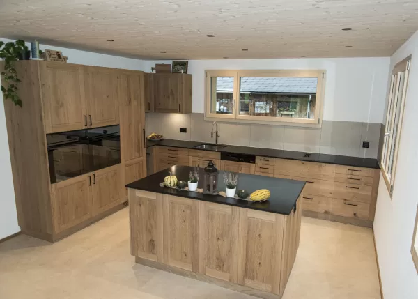 Massgefertigte Küche aus warmem Eichenholz mit zentraler Kochinsel und dunkler Arbeitsplatte. Die Kombination aus natürlichen Holzfronten, klaren Linien und modernen Einbaugeräten schafft eine zeitlose und funktionale Raumgestaltung. Grosszügige Fenster sorgen für viel Tageslicht und unterstreichen die helle, freundliche Wohnatmosphäre.