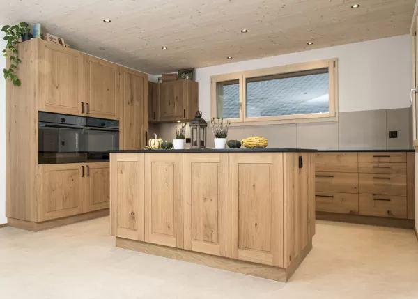 Massgefertigte Küche aus warmem Eichenholz mit zentraler Kochinsel und dunkler Arbeitsplatte. Die Kombination aus natürlichen Holzfronten, klaren Linien und modernen Einbaugeräten schafft eine zeitlose und funktionale Raumgestaltung. Grosszügige Fenster sorgen für viel Tageslicht und unterstreichen die helle, freundliche Wohnatmosphäre.