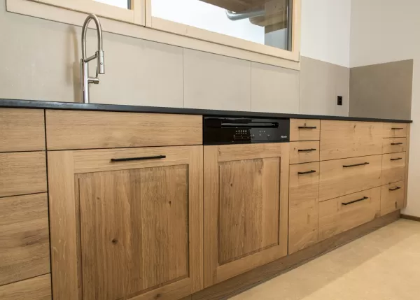 Massgefertigte Küche aus warmem Eichenholz mit zentraler Kochinsel und dunkler Arbeitsplatte. Die Kombination aus natürlichen Holzfronten, klaren Linien und modernen Einbaugeräten schafft eine zeitlose und funktionale Raumgestaltung. Grosszügige Fenster sorgen für viel Tageslicht und unterstreichen die helle, freundliche Wohnatmosphäre.