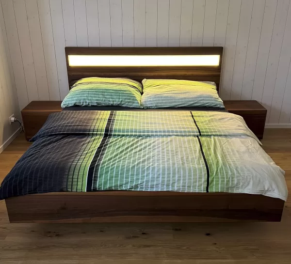 Ein modernes Doppelbett aus dunkel gemasertem Holz steht im Mittelpunkt des Schlafzimmers. Das Kopfteil ist mit einem horizontalen, warm leuchtenden Lichtstreifen versehen, der den Raum sanft ausleuchtet. Auf dem Bett liegt eine Bettwäsche in harmonischen Grün-, Blau- und Weißtönen mit einem feinen Linienmuster. Links und rechts befinden sich passende Nachttische aus dem gleichen Holz. Der Raum hat einen hellen Holzboden sowie weiss vertäfelte Wände, die eine ruhige, natürliche Atmosphäre schaffen.