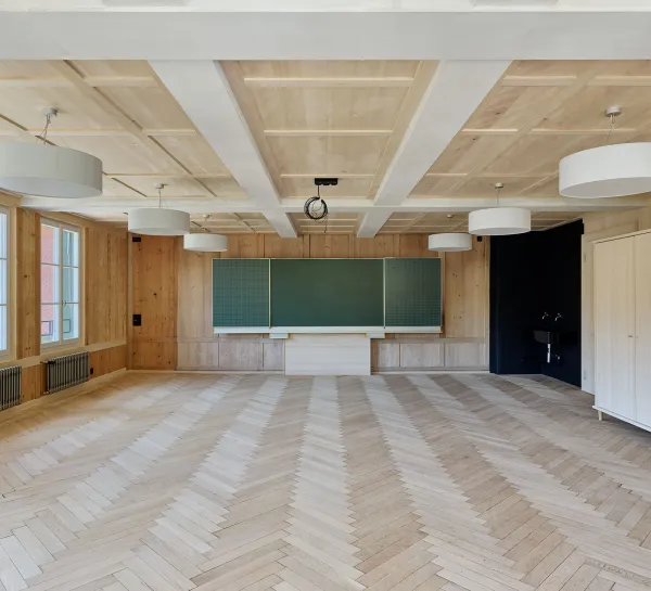 Für dieses Bild würde ich den Alt-Text so formulieren:  "Heller, mit Holz ausgekleideter Schulraum mit Fischgrätparkett, Kassettendecke, grossen Fenstern, Hängeleuchten und einer grünen Wandtafel.