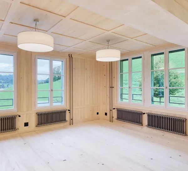 Für dieses Bild würde ich den Alt-Text so formulieren:  "Heller, mit Holz ausgekleideter Schulraum mit Fischgrätparkett, Kassettendecke, grossen Fenstern, Hängeleuchten und einer grünen Wandtafel.